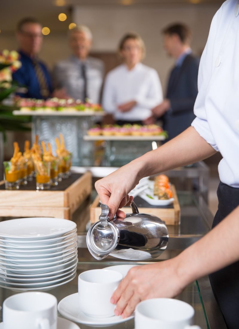 Waitress serving coffee in buffet restaurant. Group of four business people talking in background